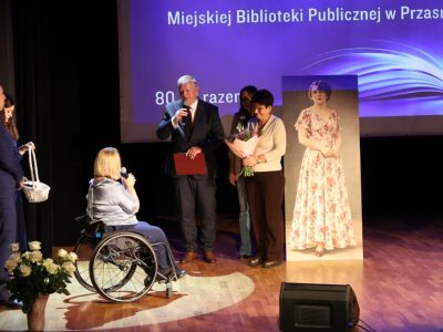 Mężczyzna w garniturze przemawia do mikrofonu, trzymając czerwoną teczkę, a obok niego stoi kobieta trzymająca bukiet kwiatów