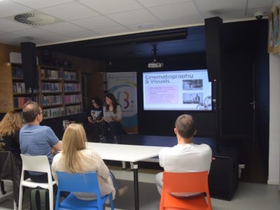 Uczestnicy Kawiarenki Językowej tyłem do obiektywu, skupieni na prezentacji multimedialnej w sali bibliotecznej. Na dużym ekranie w głębi wyświetlany jest slajd o tytule „Cinematography & Visuals” (Kinematografia i oprawa wizualna) zawierający angielski tekst oraz dwa czarno-białe kadry filmowe. Na scenie obok ekranu siedzą dwie rozmawiające kobiety prowadzące spotkanie