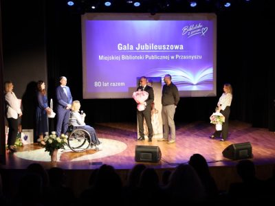 Szerokie ujęcie sceny, na której dwaj mężczyźni – jeden trzymający duży bukiet róż – przemawiają do mikrofonu, zwracając się do dyrektor biblioteki. Po obu stronach stoją pozostali goście i prowadzący, a w tle wyświetla się slajd „Gala Jubileuszowa Miejskiej Biblioteki Publicznej w Przasnyszu”.
