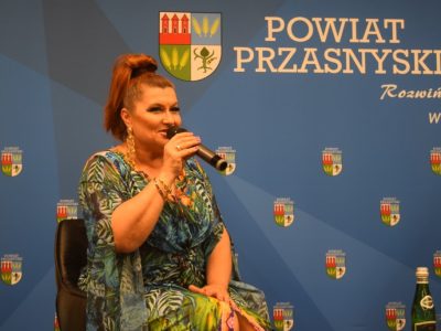 Bliskie ujęcie na uśmiechniętą Katarzynę Skrzynecką w kolorowej kreacji, która trzyma mikrofon i mówi do zgromadzonej publiczności. W tle widoczne logo Powiatu Przasnyskiego