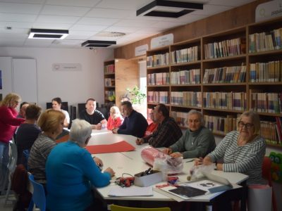 Grupa kilkunastu osób siedzi wokół połączonych białych stołów w jasnej sali bibliotecznej z regałami pełnymi książek. Na stole leżą materiały plastyczne: czerwony karton, klej na gorąco, nożyczki oraz sznurki. Uczestnicy rozmawiają ze sobą, przygotowując się do pracy manualnej