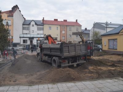Środek placu budowy, na którym stoi duża, szara ciężarówka wywrotka z pomarańczowym ramieniem dźwigu, otoczona hałdami ziemi i piasku. Za nią widoczna jest mała żółta koparka gąsienicowa. Po lewej stronie dwaj robotnicy pracują przy krawężnikach, a w głębi placu trzeci mężczyzna niesie materiały. W tle rozciąga się rząd miejskich kamienic pod zachmurzonym niebem