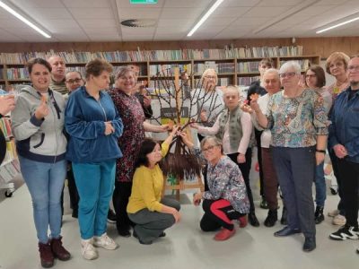 Grupa kilkunastu uczestników warsztatów stoi w bibliotece i pozuje do wspólnego zdjęcia, trzymając w dłoniach małe, jesienne liście wycięte z kolorowego papieru. W centrum grupy stoi drewniana sztaluga z brązowym drzewem wykonanym techniką makramy. W tle widoczne są wysokie, ścienne regały z literaturą