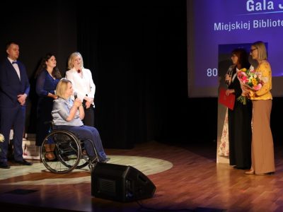 Kobieta w czarnej sukience przemawia do mikrofonu, trzymając czerwoną teczkę gratulacyjną. Obok niej stoi kobieta w żółtej bluzce z dużym, kolorowym bukietem kwiatów. Obie zwracają się do dyrektor biblioteki, obok której stoją prowadzący uroczystość