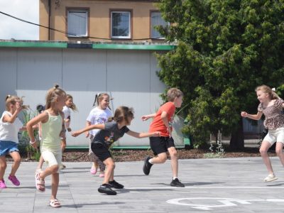 Grupa dzieci bawi się dynamicznie w berka na wybrukowanym placu przed budynkiem. W tle rosną zielone iglaki, a na pierwszym planie widoczne jest namalowane na kostce białe oznakowanie miejsca dla osób z niepełnosprawnościami