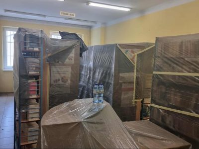 Wnętrze biblioteki przygotowane do remontu. Wysokie regały wypełnione książkami zostały szczelnie owinięte ciemną, ochronną folią malarską i zabezpieczone taśmą. Na pierwszym planie, na również zabezpieczonym folią stoliku, stoją dwie plastikowe butelki z wodą. W tle widać żółte ściany oraz okno, a pod sufitem tabliczkę z napisem „LITERATURA PIĘKNA”