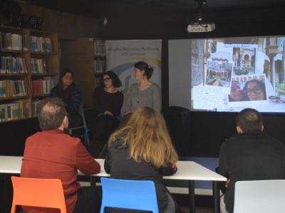 Uczestnicy siedzący przodem do prelegentek obserwują kawiarenkę językową prowadzoną przez trzy młode kobiety. Na ekranie wyświetlona jest prezentacja, na której widać kolaż ze zdjęć, najprawdopodobniej wspomnienia z wycieczki