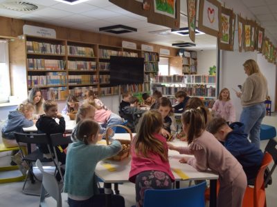 Dzieci siedzą w bibliotece przy połączonych białych stołach, rysując i kolorując. W tle widać wysokie drewniane regały z książkami, a z sufitu zwisają sznurki z przypiętymi pracami plastycznymi. Dwie dorosłe opiekunki nadzorują pracę grupy