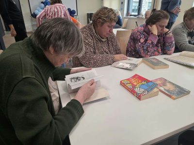Kobieta w ciemnozielonym swetrze czyta otwartą książkę, obok na stole leżą inne publikacje, m.in. „Klątwa złamanej nogi”. Obok siedzą dwie kobiety skupione na lekturze