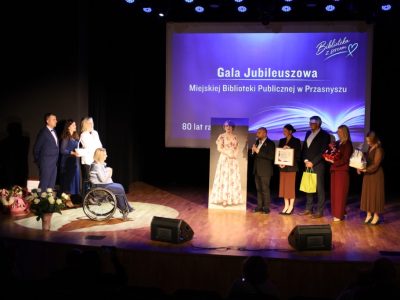 Szerokie ujęcie sceny, na której grupa pięciorga delegatów z dyplomem, torbą i zapakowanymi upominkami przemawia do dyrektor biblioteki