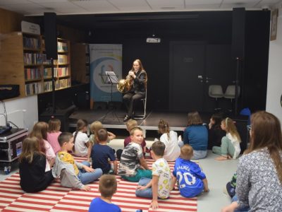 Dziewczyna w ciemnym garniturze gra na waltorni, siedząc przy pulpicie na małej scenie w bibliotece. Przed nią, na pasiastym dywanie, siedzi grupa dzieci słuchających koncertu