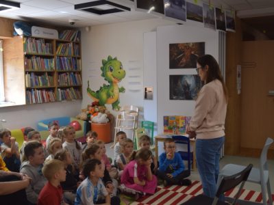 Kobieta w okularach i jasnej bluzie stoi przed grupą przedszkolaków siedzących na pasiastym dywanie w bibliotece i rozmawia z nimi. Dzieci z uwagą słuchają i patrzą w jej stronę. W tle widoczny jest kącik dziecięcy z regałami pełnymi książek, dużą naklejką przedstawiającą zielonego smoka na ścianie oraz niskimi, kolorowymi krzesełkami