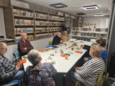 Uczestnicy Dyskusyjnego Klubu Książki zgromadzeni wokół jasnego stołu w bibliotece. Jeden z mężczyzn trzyma w rękach pomarańczową książkę z tytułem „James”. W tle regały biblioteczne oraz napis „JESIEN”