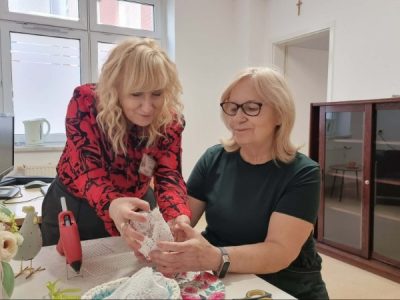 W centralnej części zdjęcia widać bibliotekarkę, która pomaga czytelniczce stworzyć wielkanocną ozdobę. Za nimi po lewej stronie stoi komputer dla użytkowników. Po prawej stronie zdjęcia znajduje się brązowa witryna.