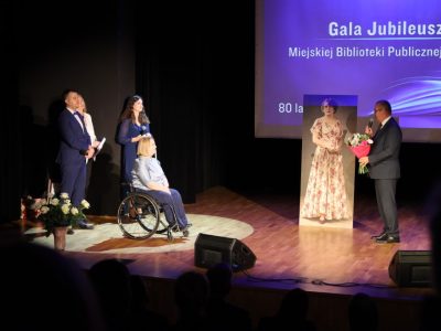 Mężczyzna w garniturze przemawia do mikrofonu, trzymając oprawiony dyplom przed gospodarzami wydarzenia. W tle znajduje się niebieski ekran z napisem „Gala Jubileuszowa Miejskiej Biblioteki Publicznej w Przasnyszu – 80 lat razem”