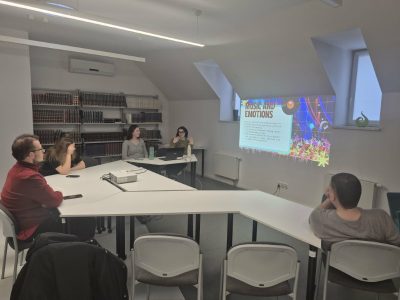 Prezentacja w oddziale dla dorosłych podczas kawiarenki językowej. Na ścianie wyświetlany jest slajd w języku angielskim „MUSIC AND EMOTIONS”. Wolontariuszka w zielonym swetrze siedzi przy laptopie i gestykuluje, prowadząc dyskusję z uczestnikami zgromadzonymi wokół stołu