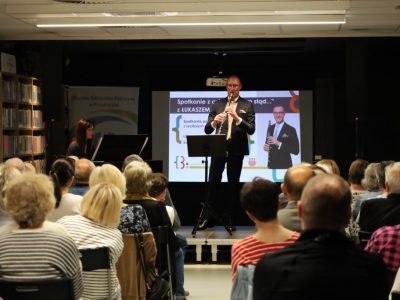 Łukasz Motyka gra na klarnecie na podwyższeniu, w tle widoczna pianistka oraz ekran projekcyjny. Na pierwszym planie tyłem do obiektywu siedzi publiczność zapełniająca salę biblioteczną