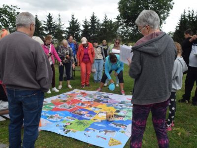 Grupa dorosłych i dzieci stoi w kręgu na trawie wokół rozłożonej na ziemi wielkoformatowej, kolorowej gry planszowej z ilustracjami. Jedna z kobiet w niebieskiej bluzie pochyla się nad planszą, przesuwając element gry, podczas gdy pozostali uczestnicy, w tym kobieta z dokumentami w ręku, uważnie obserwują jej ruch