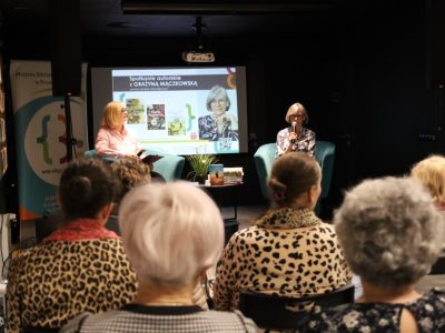 Spotkanie autorskie z pisarką Grażyną Mączkowską w bibliotece. Autorka siedzi w fotelu na scenie i rozmawia z prowadzącą, przed nimi znajduje się publiczność. W tle widoczny jest ekran z napisem "Spotkanie autorskie z Grażyną Mączkowską" oraz okładkami jej książek