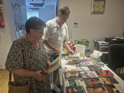Kobieta w kwiecistej bluzce oraz mężczyzna w jasnej koszuli stoją przy białym stole w bibliotece, przeglądając i wybierając książki. Na blacie rozłożonych jest kilkadziesiąt różnych powieści oraz stojak z informacjami. W tle, na ścianie nad nimi, wisi klasyczny zegar wskazówkowy