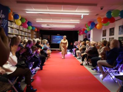 Uśmiechnięta seniorka prezentuje elegancką, beżową dzianinową sukienkę i torebkę, idąc po czerwonym dywanie. Z obu stron otaczają ją zgromadzeni goście bijący brawo