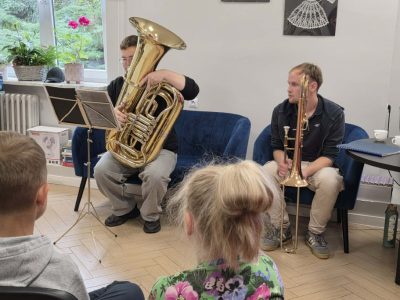 Dwóch młodych mężczyzn siedzi na niebieskich kanapach w sali i gra na instrumentach – jeden na tubie przy nutach na stojaku, a drugi trzyma puzon. W pierwszym planie widać tył głów słuchających ich dzieci