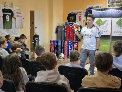 Widok z perspektywy siedzących na krzesłach dzieci na prowadzącego w koszulce Realu Madryt, który opowiada i gestykuluje. W tle widoczne są eksponowane na wieszakach i ścianie koszulki piłkarskie