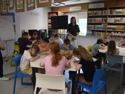 Grupa dzieci siedzi przy białych, modułowych stołach w jasnej sali bibliotecznej i wykonuje prace plastyczne. Dzieci pochylają się nad stołami, rysując pisakami i kredkami. Pośrodku jednego ze stołów stoi wiklinowy koszyk. W tle stoją dwie dorosłe kobiety, które obserwują pracę uczestników. Z prawej strony widoczne są wysokie regały ścienne z lekturami i książkami dla młodzieży