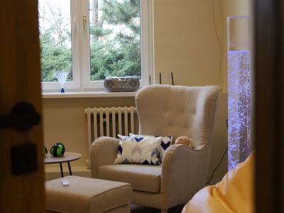 Widok przez otwarte drzwi na pokój wyciszeń w Filii nr 1. W centrum znajduje się beżowy fotel z poduszką i królikiem obciążeniowym, obok kolumna świetlna i okno z widokiem na drzewa iglaste. Na parapecie stoi radioodtwarzacz