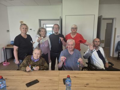 Siedmiu uczestników zajęć rękodzielniczych – kobiety i mężczyźni (w tym osoba na wózku inwalidzkim) – pozuje razem do pamiątkowej fotografii. Każdy z dumą trzyma w dłoni i prezentuje ukończony kolorowy brelok ze sznurka