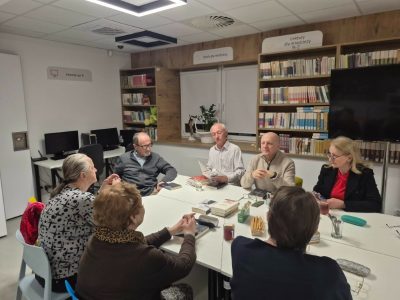 Grupa siedmiu dorosłych czytelników siedzi przy stole podczas spotkania Dyskusyjnego Klubu Książki. Jeden z uczestników sprawia wrażenie, jakby odczytywał fragment książki.