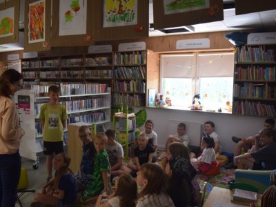 Uczestnicy warsztatów siedzą na kolorowym dywanie i pufach w strefie dla dzieci w bibliotece. Po lewej stronie stoi uśmiechnięta prowadząca, która rozmawia z jednym z chłopców. Nad regałami z książkami wiszą dziecięce rysunki, a w głębi znajduje się jasne okno z zabawkami na parapecie