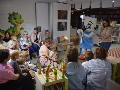 Grupa małych dzieci wraz z opiekunami siedzi w kręgu w kąciku dziecięcym w bibliotece. Wszyscy z uwagą patrzą na postać w dużym kostiumie maskotki Kici Koci, która stoi przed nimi w niebieskiej sukience i białych rękawiczkach. Obok maskotki stoi kobieta trzymająca otwartą książkę. Na ścianie po lewej stronie widnieje duża naklejka z zielonym smokiem, a na podłodze leży kolorowa mata z zabawkami