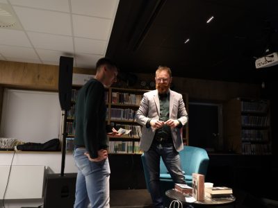 Krzysztof Piersa stoi w sali bibliotecznej i rozmawia z mężczyzną trzymającym banknot. Przed nimi na stoliku leżą egzemplarze książek autora