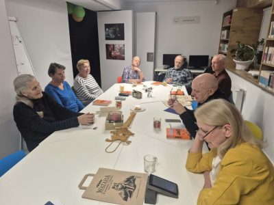 czestnicy spotkania klubowego siedzący przy stole rozmawiają i słuchają dyskusji. Na pierwszym planie widać kobietę w żółtej żakiecie oraz torbę z logo „Wataha”