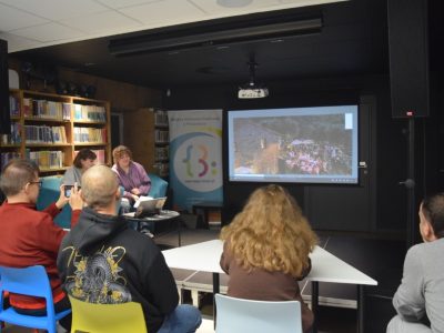 Ujęcie od tyłu na widownię zgromadzoną przy stołach podczas spotkania w bibliotece. Jeden z uczestników robi zdjęcie telefonem, a z przodu dwie prelegentki prowadzą prezentację