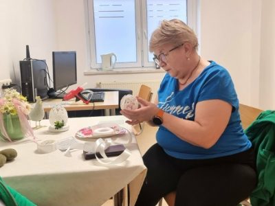 Kobieta w niebieskiej bluzce siedzi przy stole, gdzie tworzy wielkanocne ozdoby z akrylowych jajek. Na stole przed nią znajdują się materiały potrzebne do stworzenia pracy.
