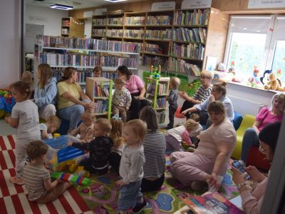 Swobodna zabawa dzieci i opiekunów po spotkaniu w bibliotece. Maluchy siedzą na kolorowym dywanie, układają plastikowe klocki z dużego pojemnika i bawią się zabawkami edukacyjnymi. Dorośli siedzą obok nich na podłodze lub na niskich, kolorowych pufach. W tle widoczne są regały z literaturą dziecięcą oraz duże okno, na którym stoją pluszowe maskotki