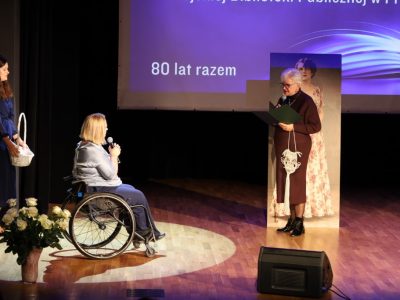 Kobieta w okularach i ciemnej sukience z ozdobną makramą czyta życzenia z teczki do mikrofonu, stojąc naprzeciwko dyrektor biblioteki