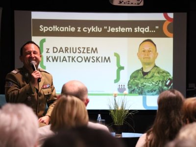 Bliskie ujęcie z perspektywy widowni na uśmiechniętego generała Dariusza Kwiatkowskiego trzymającego mikrofon. W tle znajduje się ekran prezentacji z napisem „Spotkanie z cyklu Jestem stąd… z DARIUSZEM KWIATKOWSKIM”