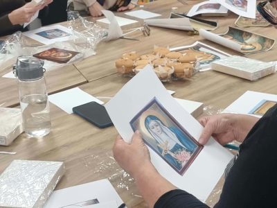 Kobieta w okularach trzyma w dłoniach arkusz papieru z kolorowym wizerunkiem świętej postaci, który służy jako wzór do wykonania ikony. Na stole obok leżą deseczki oraz poczęstunek