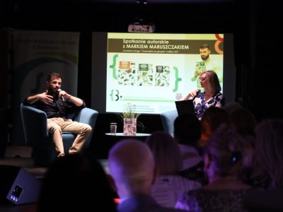 Przebieg spotkania autorskiego z Markiem Maruszczakiem z perspektywy publiczności. Na oświetlonej scenie autor i prowadząca rozmawiają przez mikrofony, siedząc naprzeciwko siebie w fotelach. Między nimi stoi mały stolik z książką i szklanką wody. W tle jasny ekran z plakatem wydarzenia. Pierwszy plan stanowią zaciemnione sylwetki i tyły głów siedzących widzów