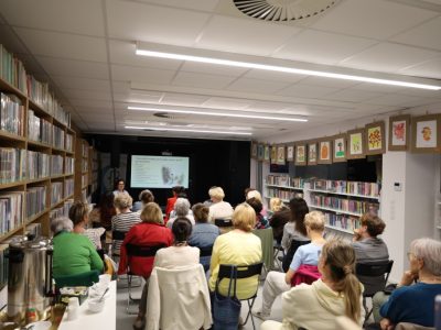 Perspektywa z boku sali bibliotecznej, ukazująca liczną grupę dorosłych osób słuchających wykładu. Nad regałami po prawej stronie wiszą oprawione rysunki dziecięce. Na pierwszym planie po lewej stronie stoi stolik z cateringiem, na którym umieszczono duży, metalowy termos gastronomiczny na napoje oraz białe filiżanki