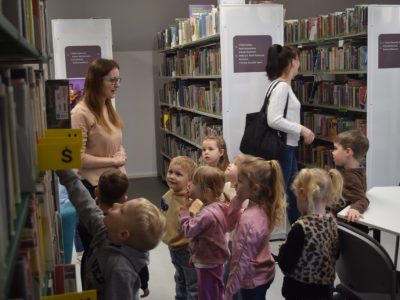 Grupa przedszkolaków spaceruje między regałami w bibliotece pod opieką bibliotekarki. Jedno z dzieci wyciąga rączkę w stronę książek na półce. Bibliotekarka uśmiecha się i opowiada o zbiorach