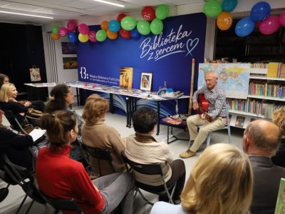 Widok z góry na kameralne spotkanie autorskie w bibliotece. Podróżnik Andrzej Ciuba siedzi przy regale z książkami i gra na gitarze klasycznej, a przed nim siedzi na krzesłach wpatrzona publiczność. Na stołach w tle wyłożone są pamiątki i zdjęcia z wypraw
