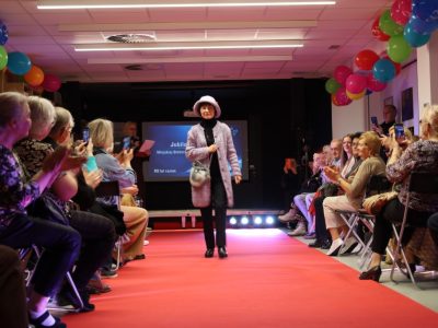 Modelka w lawendowym, miękkim płaszczu, dopasowanym kapeluszu i z torebką idzie po czerwonym dywanie wzdłuż rzędów krzeseł z publicznością