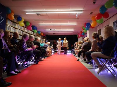 Dwie seniorki prezentują dzianinowe stroje na czerwonym dywanie w bibliotece. Publiczność siedząca po obu stronach wybiegu bije brawo i robi zdjęcia telefonami