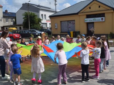 Grupa dzieci stoi w kole na wybrukowanym podwórku i trzyma tęczową chustę animacyjną. Po lewej stronie stoi tyłem dorosła kobieta (opiekunka) pomagająca w zabawie. W tle widoczny jest parterowy, żółty budynek z tablicą „Dom Harcerza” oraz zaparkowane samochody