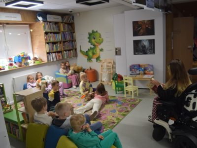 Grupa dzieci siedzi na kolorowym dywanie i pufach w strefie dziecięcej biblioteki, słuchając czytanej opowieści. Niektóre dzieci trzymają pluszowe zabawki. Po prawej stronie widoczna jest Sylwia Błach, która trzyma otwartą książkę i czyta ją grupie. W tle regały z książkami oraz dekoracja z zielonym smokiem na ścianie