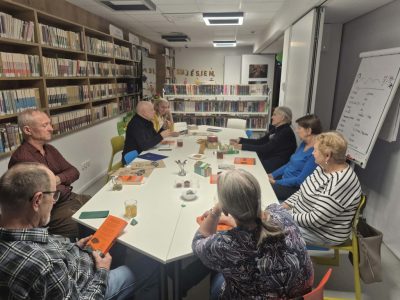 Ujęcie z boku na uczestników Dyskusyjnego Klubu Książki siedzących przy podłużnym stole w bibliotece. Obok nich stoi flipchart z zapiskami i rysunkami